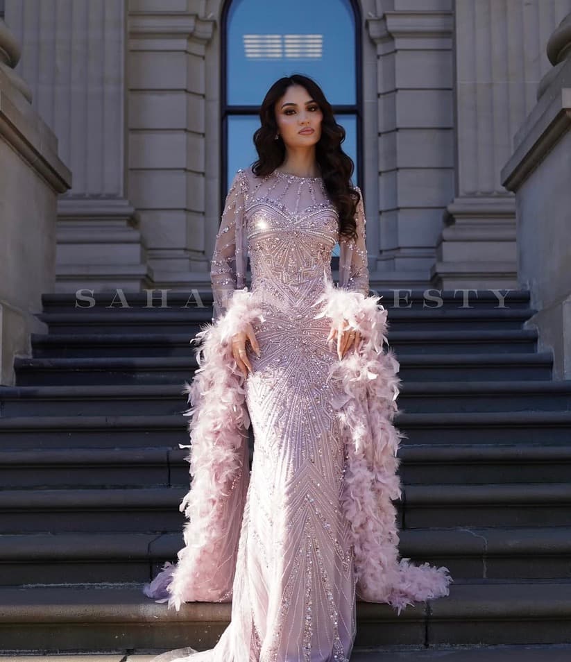 Model in a blush crystal modest gown with feather sleeves on stone steps.