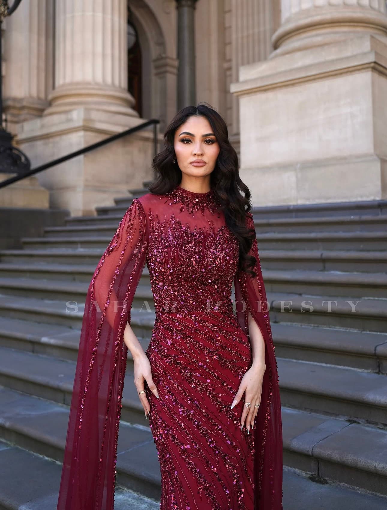Close view of a burgundy beaded modest gown with cape sleeves.