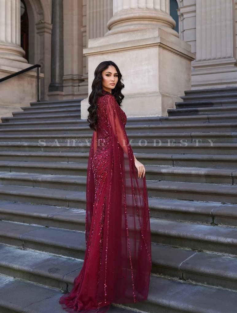 Burgundy beaded modest cape gown shown from the side on stone steps.