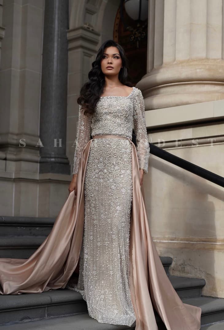 Model in a champagne column modest gown with a satin overskirt beside a stone column.