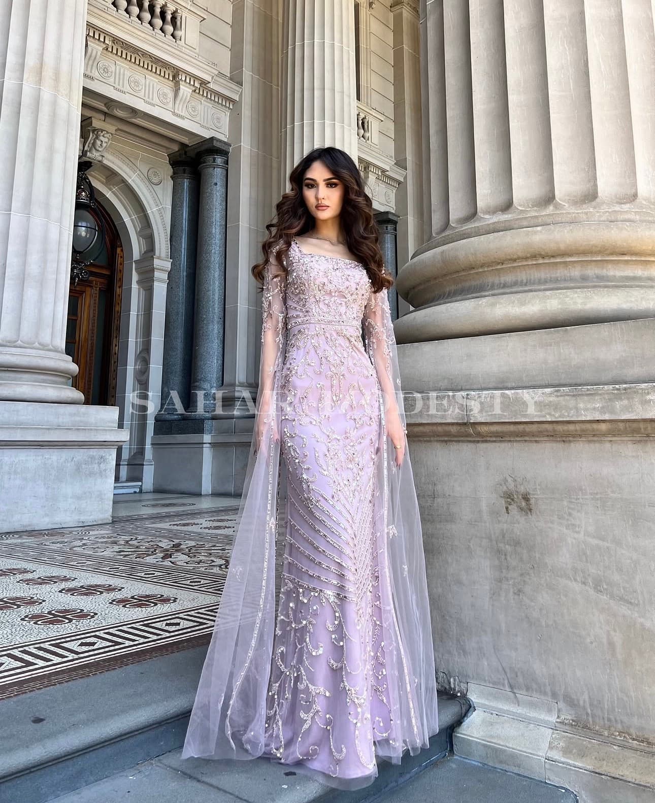 Model in a lilac beaded modest gown with a sheer overlay beside stone columns.