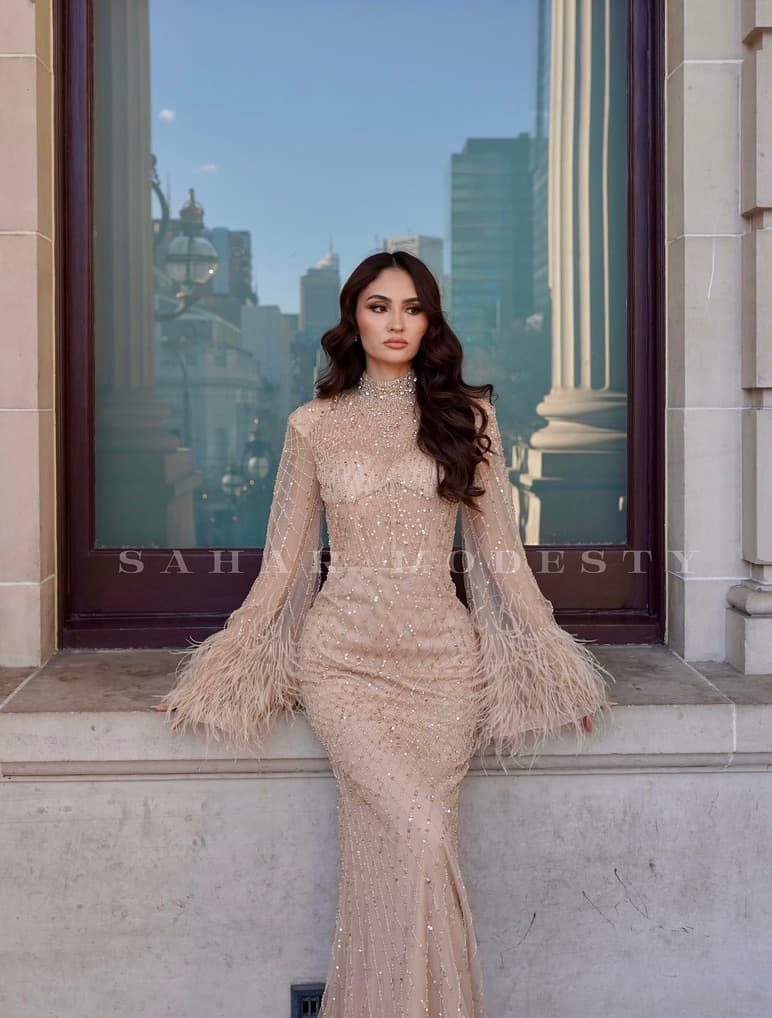Model in a nude beaded modest gown with full feather cape sleeves.