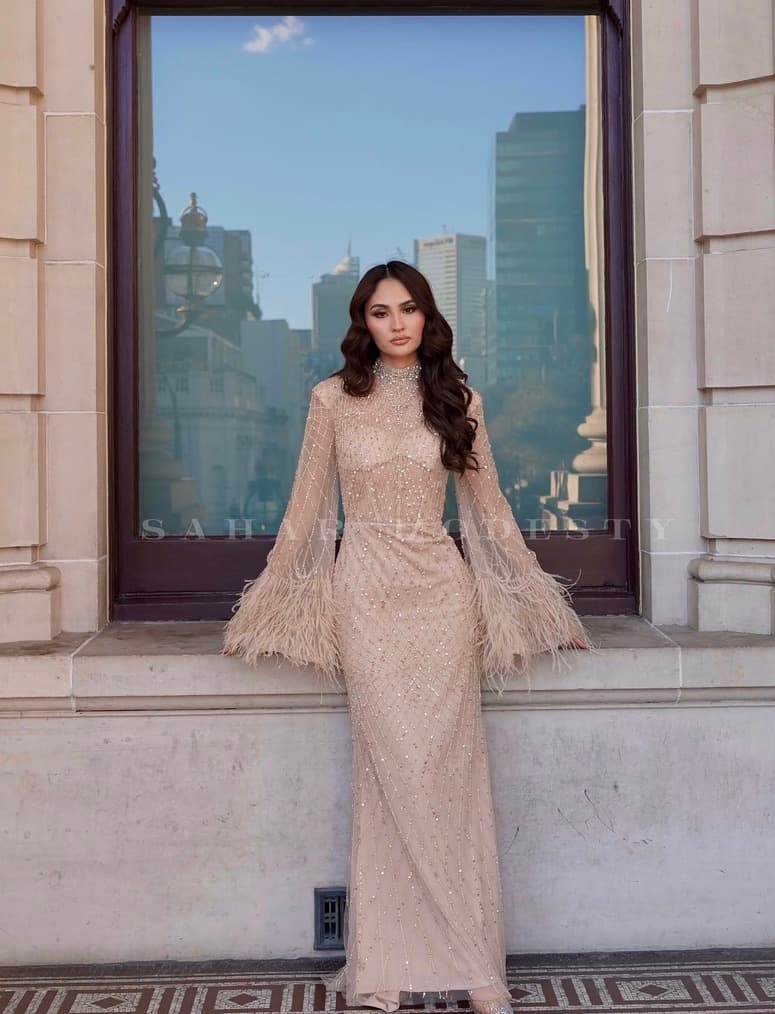 Nude beaded modest feather sleeve gown in front of a reflective window.